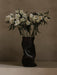 The black, sculptural Audo Collapse Vase sits on a light surface, holding a bouquet of white flowers against a taupe wall.