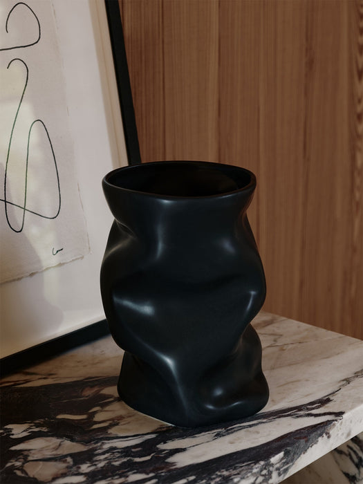 A close-up of the black ceramic Collapse Vase on a marble Plinth Bridge, styled with framed art against a wood wall.