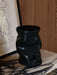 A close-up of the black ceramic Collapse Vase on a marble Plinth Bridge, styled with framed art against a wood wall.
