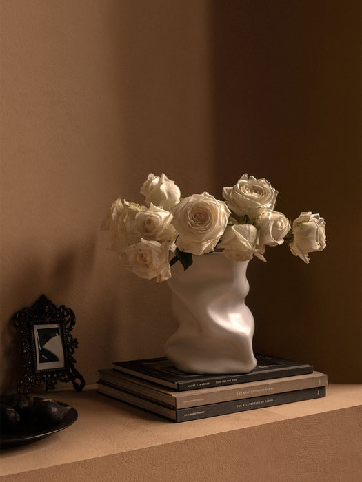 A white, sculptural Collapse Vase holds white roses, sitting on a stack of books on a shelf next to a picture frame & fruit.