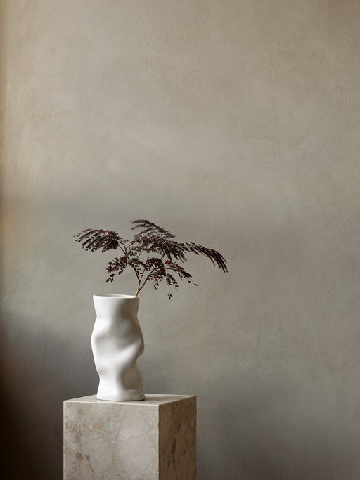 The sculptural white Collapse Vase holding a dark-leafed branch, styled on a square marble Plinth Pedestal.