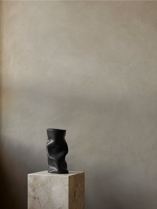 A matte black, sculptural Collapse Vase displayed on a cubic, veined marble Plinth Pedestal against a neutral, textured wall.