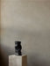 A matte black, sculptural Collapse Vase displayed on a cubic, veined marble Plinth Pedestal against a neutral, textured wall.