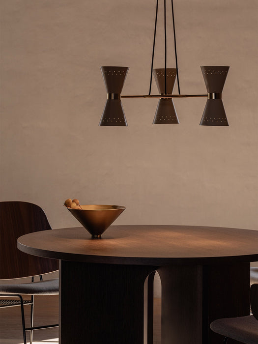 The brass and dark metal Collector Chandelier, 3 hangs over a dark wood Androgyne Dining Table with a decorative brass bowl.