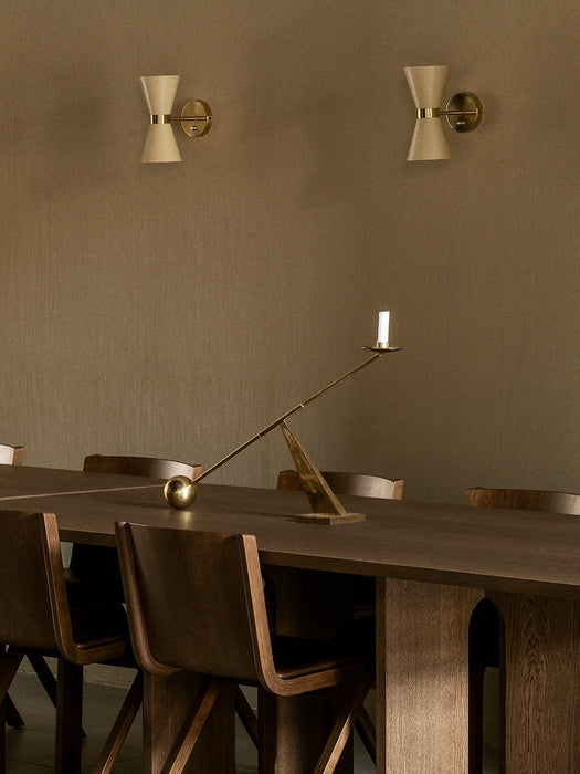 Two cream and brass Collector Wall Lamps on a beige wall over a dark wood dining table with chairs and brass candleholders.