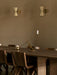 Two cream and brass Collector Wall Lamps on a beige wall over a dark wood dining table with chairs and brass candleholders.