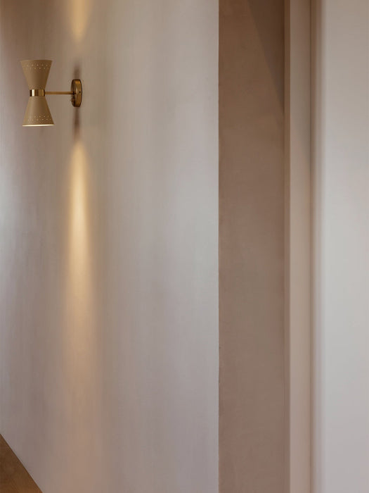 A cream and brass Collector Wall Lamp mounted on a pale wall, casting warm light upwards and downwards in a hallway setting.