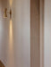 A cream and brass Collector Wall Lamp mounted on a pale wall, casting warm light upwards and downwards in a hallway setting.