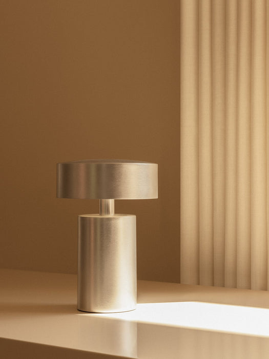 A close-up of the Column Table Lamp in brushed aluminium, sitting on a pale surface with a beam of light in a warm setting.