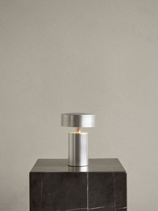 The Column Table Lamp, Portable in a brushed aluminium finish, switched on and placed on a tall, dark marble plinth.