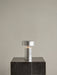 The Column Table Lamp, Portable in a brushed aluminium finish, switched on and placed on a tall, dark marble plinth.