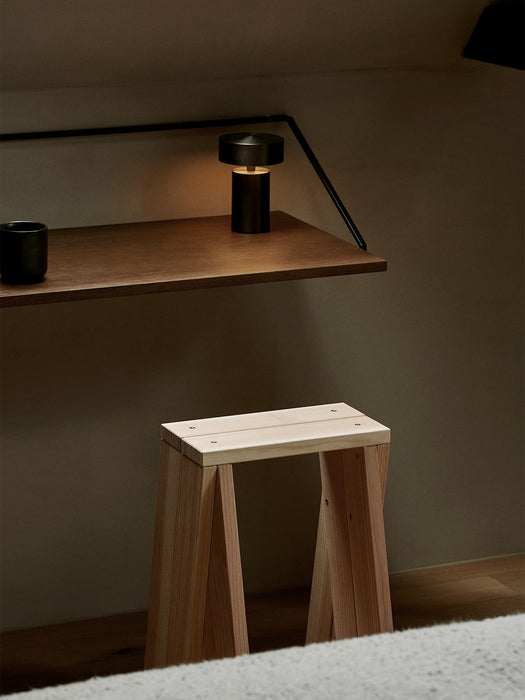 The black Column Table Lamp glowing on a dark wood Rail Shelf, positioned above a natural wood Ishinomaki Stool in a corner.