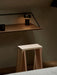 The black Column Table Lamp glowing on a dark wood Rail Shelf, positioned above a natural wood Ishinomaki Stool in a corner.