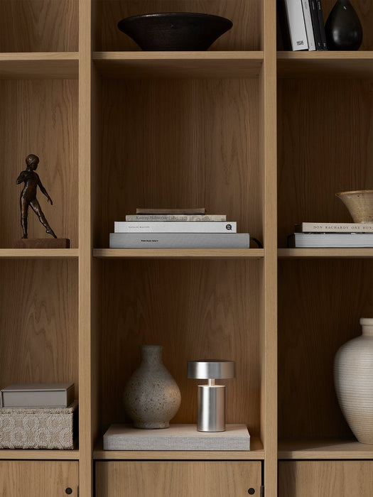 The Brushed Aluminium Column Table Lamp, Portable, sits on a book in a wooden bookshelf, styled with vases and sculptures.