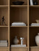 The Brushed Aluminium Column Table Lamp, Portable, sits on a book in a wooden bookshelf, styled with vases and sculptures.