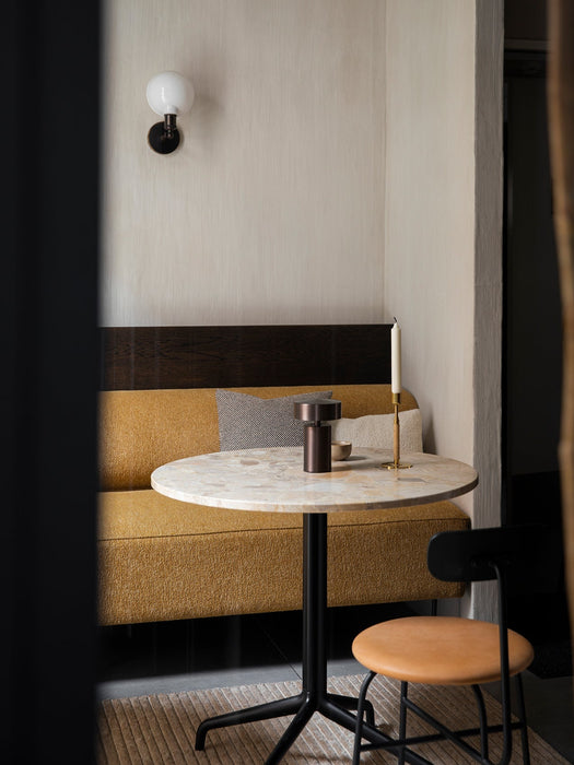 The Column Table Lamp on a round marble table in a cosy restaurant corner with a mustard-coloured bench and a black chair.