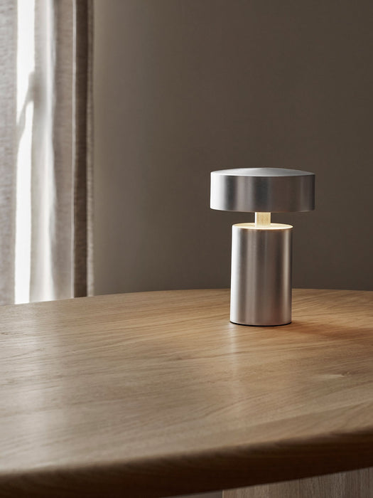 A portable Column Table Lamp with a brushed aluminium finish on a natural oak desk, its warm light reflecting on the wood.