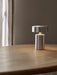 A portable Column Table Lamp with a brushed aluminium finish on a natural oak desk, its warm light reflecting on the wood.
