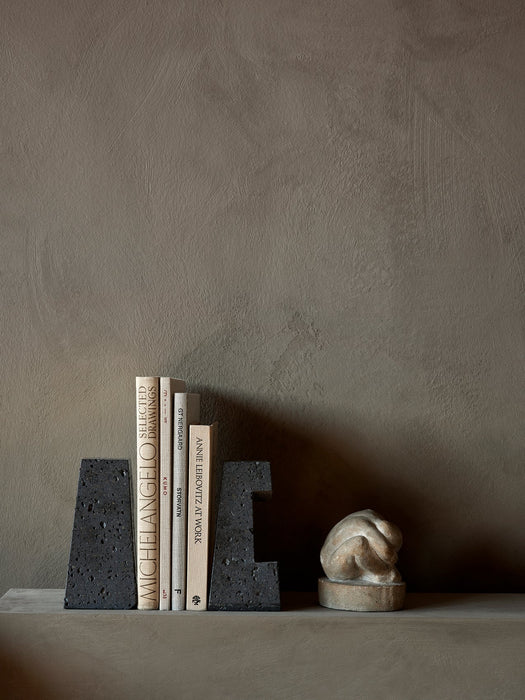 A pair of black, textured Converge Book Ends holding four books upright on a shelf, next to a small, pale abstract sculpture.