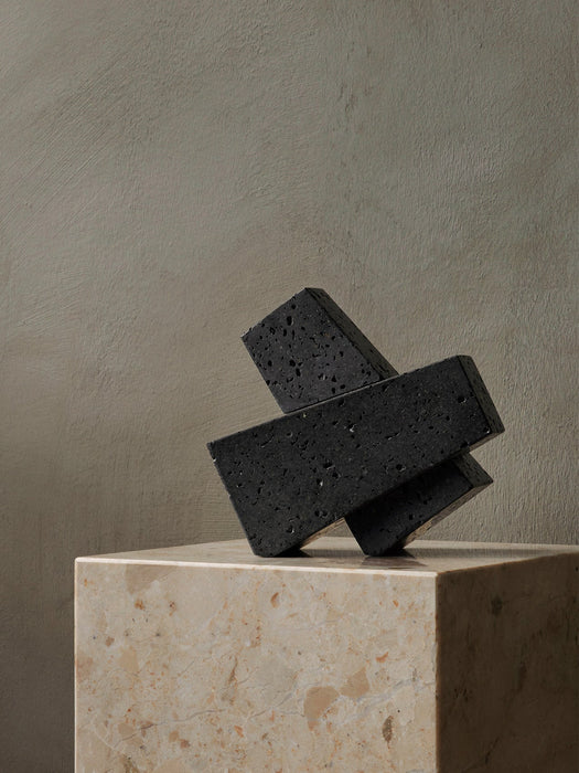 The black, porous Converge Book End, formed of three interlocking blocks, sits sculpturally atop a beige marble plinth.