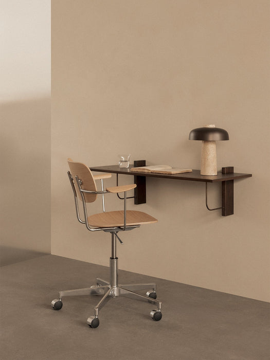Angled view of the dark wood Corbel Desk mounted on a beige wall, with a light wood Co Task Chair and a Reverse Table Lamp.