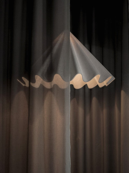 An artistic shot of the white Dancing Pendant Lamp, its glowing, pleated shade partially veiled by a sheer dark curtain.