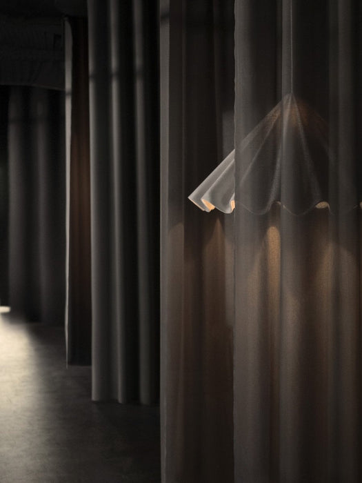 The white Dancing Pendant Lamp is seen partially obscured by a dark, sheer curtain, casting a warm glow in a dimly lit room.