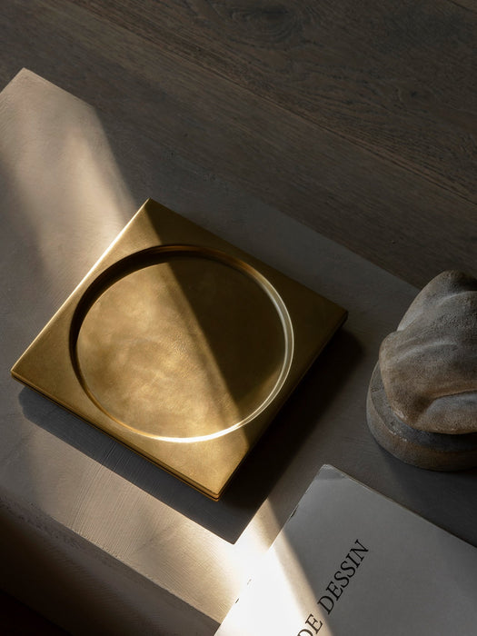 The square brass Divot Tray sits on a textured grey surface, illuminated by a sliver of sunlight next to a stone sculpture.