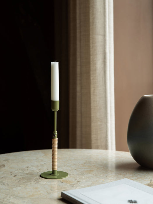A Duca Candle Holder in olive green with a rattan-wrapped stem holds a white candle on a marble tabletop in a warm setting.