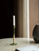 A Duca Candle Holder in olive green with a rattan-wrapped stem holds a white candle on a marble tabletop in a warm setting.