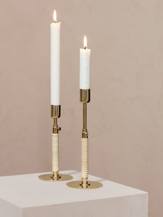 A pair of brass Duca Candleholders with rattan-wrapped stems holding lit white taper candles against a muted pink wall.