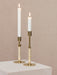 A pair of brass Duca Candleholders with rattan-wrapped stems holding lit white taper candles against a muted pink wall.