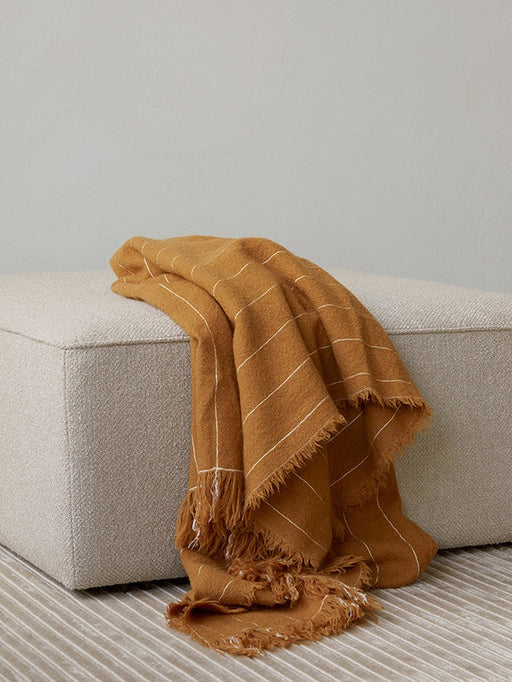 A close-up of a mustard-coloured Battus Throw with white stripes draped over a light grey bouclé Eave Modular Pouf on a rug.