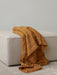 A close-up of a mustard-coloured Battus Throw with white stripes draped over a light grey bouclé Eave Modular Pouf on a rug.