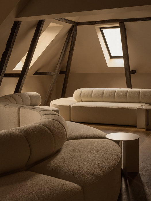 The cream Eave Seamline Modular Sofa and Androgyne Side Table in a warmly lit attic room with exposed dark wooden beams.
