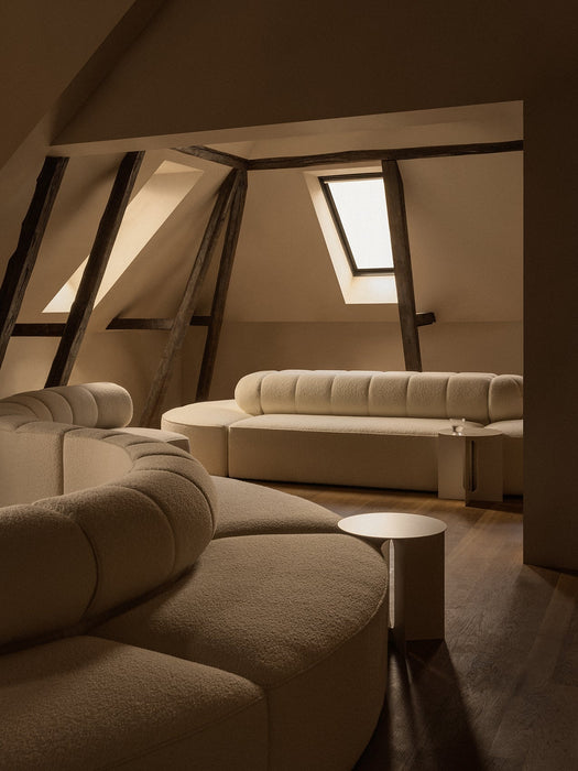 A cream Eave Seamline Modular Sofa and Androgyne Side Table in an attic room with dark wooden beams, lit by a skylight.