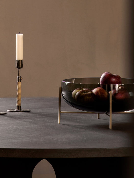 The smoked glass Echasse Bowl, filled with red apples, sits on a brass stand on a dark table next to a candlestick.