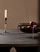 The smoked glass Echasse Bowl, filled with red apples, sits on a brass stand on a dark table next to a candlestick.