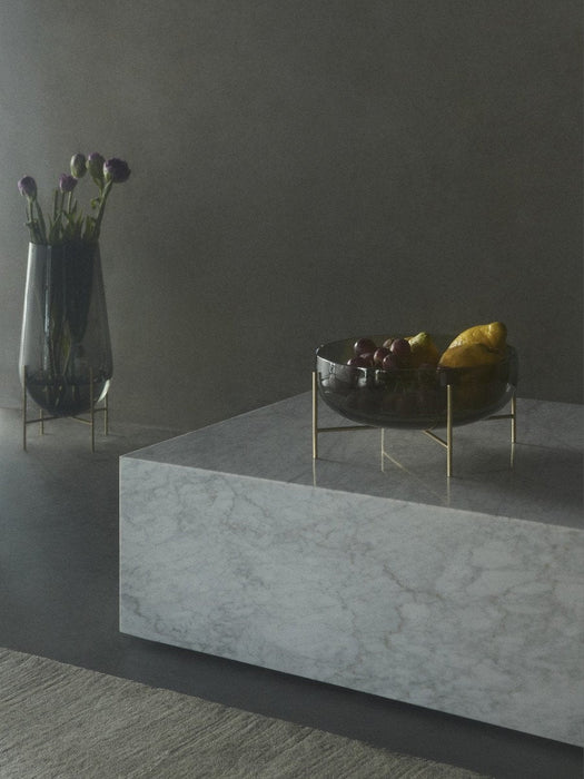 An Echasse Bowl with fruit sits on a marble Plinth, next to an Echasse Vase with purple flowers in a minimalist room.