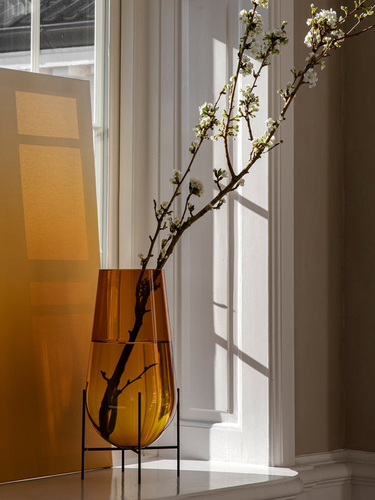 Echasse Vase in amber glass with a black stand, holding a flowering branch, bathed in sunlight on a windowsill.