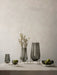 A collection of Audo CPH's Echasse vases and bowls in smoked glass on brass stands, styled with fruit and dried botanicals.