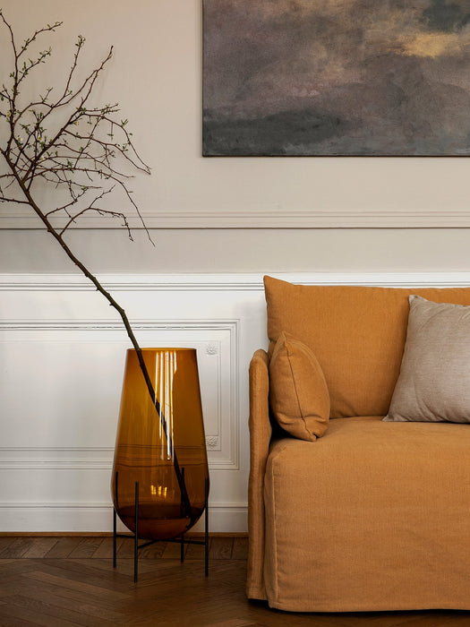 The Échasse Vase in amber glass, holding a budding branch, sits on a dark wood floor next to a mustard-coloured sofa.