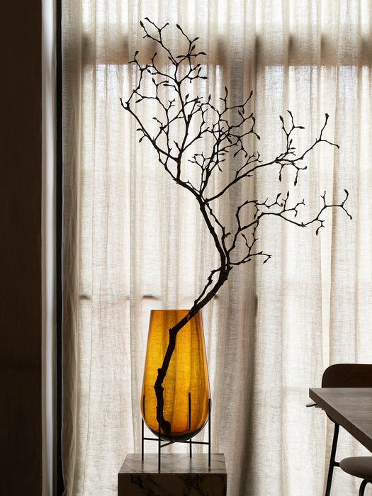 The Hasse Vase in amber glass, holding a decorative branch, is displayed on a marble Plinth in front of a sunlit window.