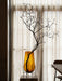 The Hasse Vase in amber glass, holding a decorative branch, is displayed on a marble Plinth in front of a sunlit window.