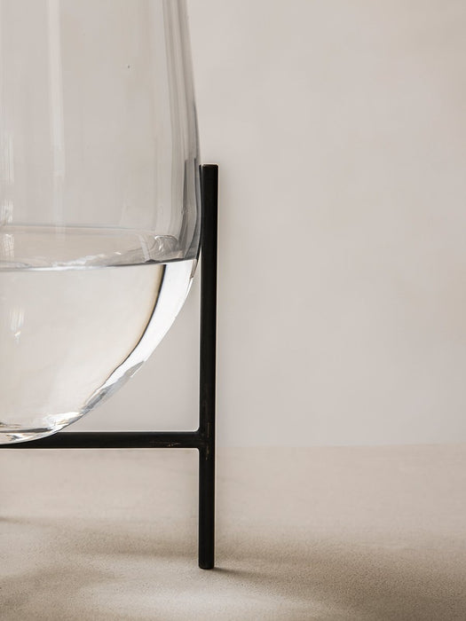 A close-up of the clear glass Echasse Vase, filled with water and cradled in its black metal stand against a neutral wall.
