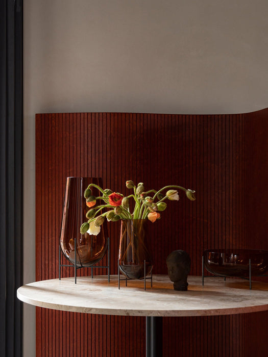 A close-up of the Hasse Vases and Bowl on a marble Column Table. Vases hold poppies in front of a reddish-brown wood panel.
