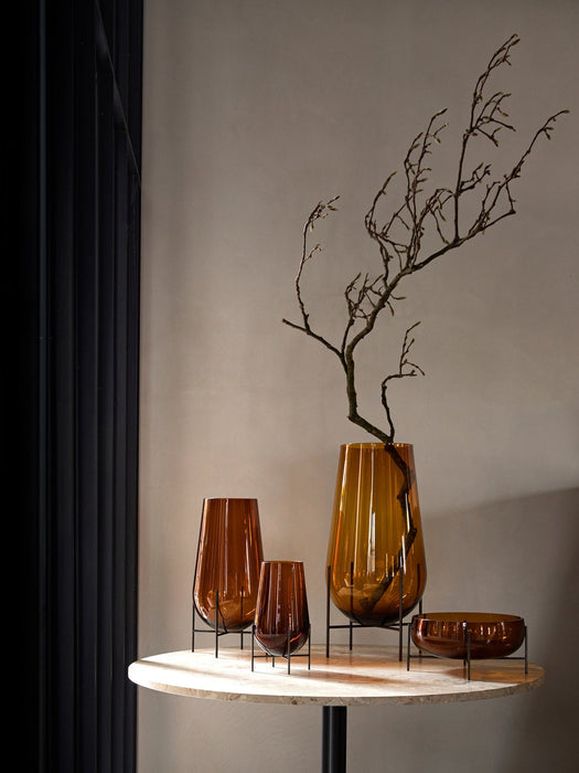 A collection of amber glass Echasse Vases and an Echasse Bowl by Audo CPH, styled on a light marble table with a branch.