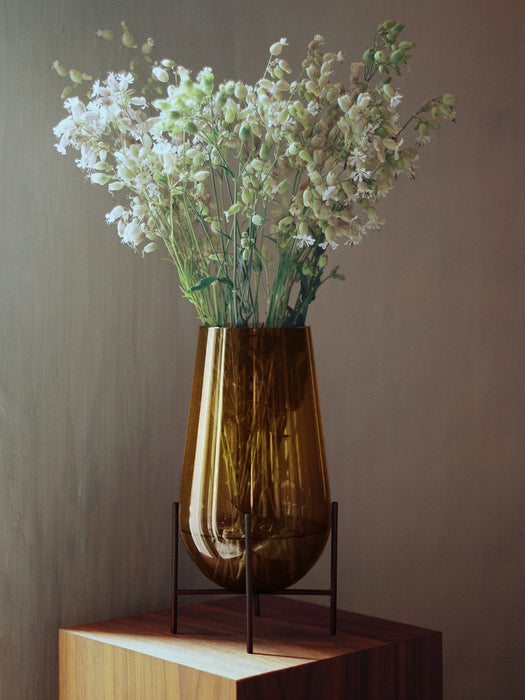 The medium Hasse Vase in amber glass, held by a black metal stand, on a wooden surface filled with white flowers.