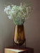 The medium Hasse Vase in amber glass, held by a black metal stand, on a wooden surface filled with white flowers.