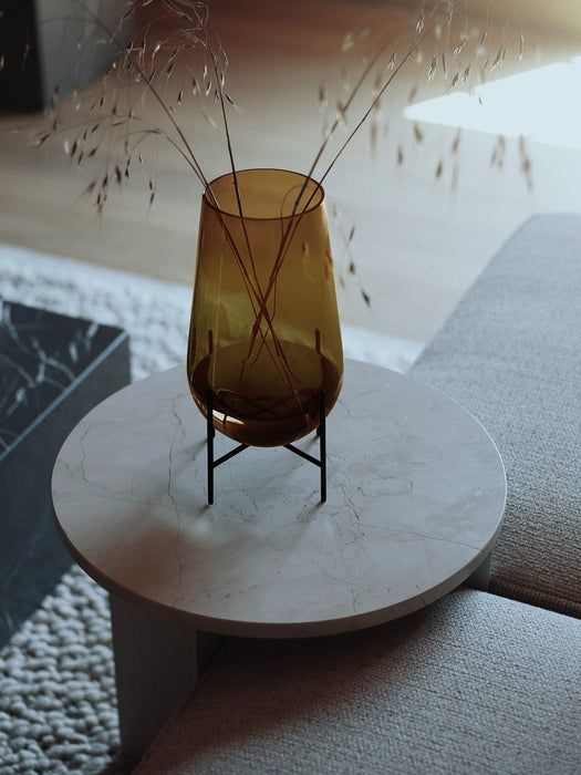 A small Hasse Vase in amber glass with dried grasses, placed on a light marble Androgyne Side Table next to a textured sofa.